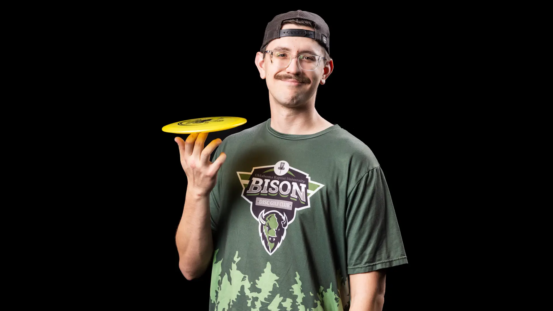 A disc golf player wearing glasses and a backwards cap balances a yellow disc on his fingertips, smiling in a green Bison Disc Golf Club shirt.