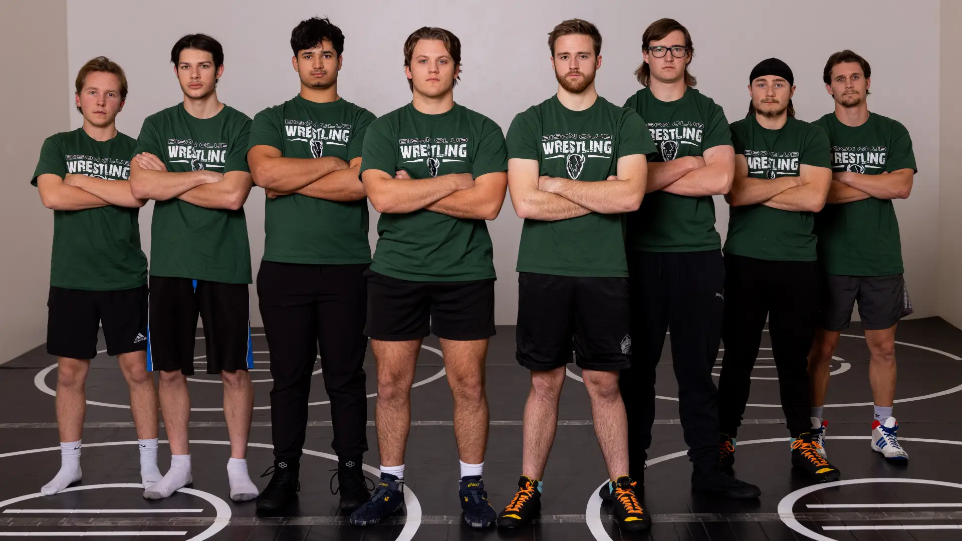 Eight members of the Wrestling Club stand side by side with arms crossed, wearing green Bison Club Wrestling t-shirts.